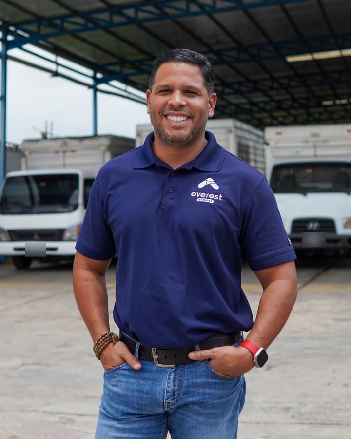 Aramis Monge sonriendo, de pie en el patio de operaciones, con camiones detrás